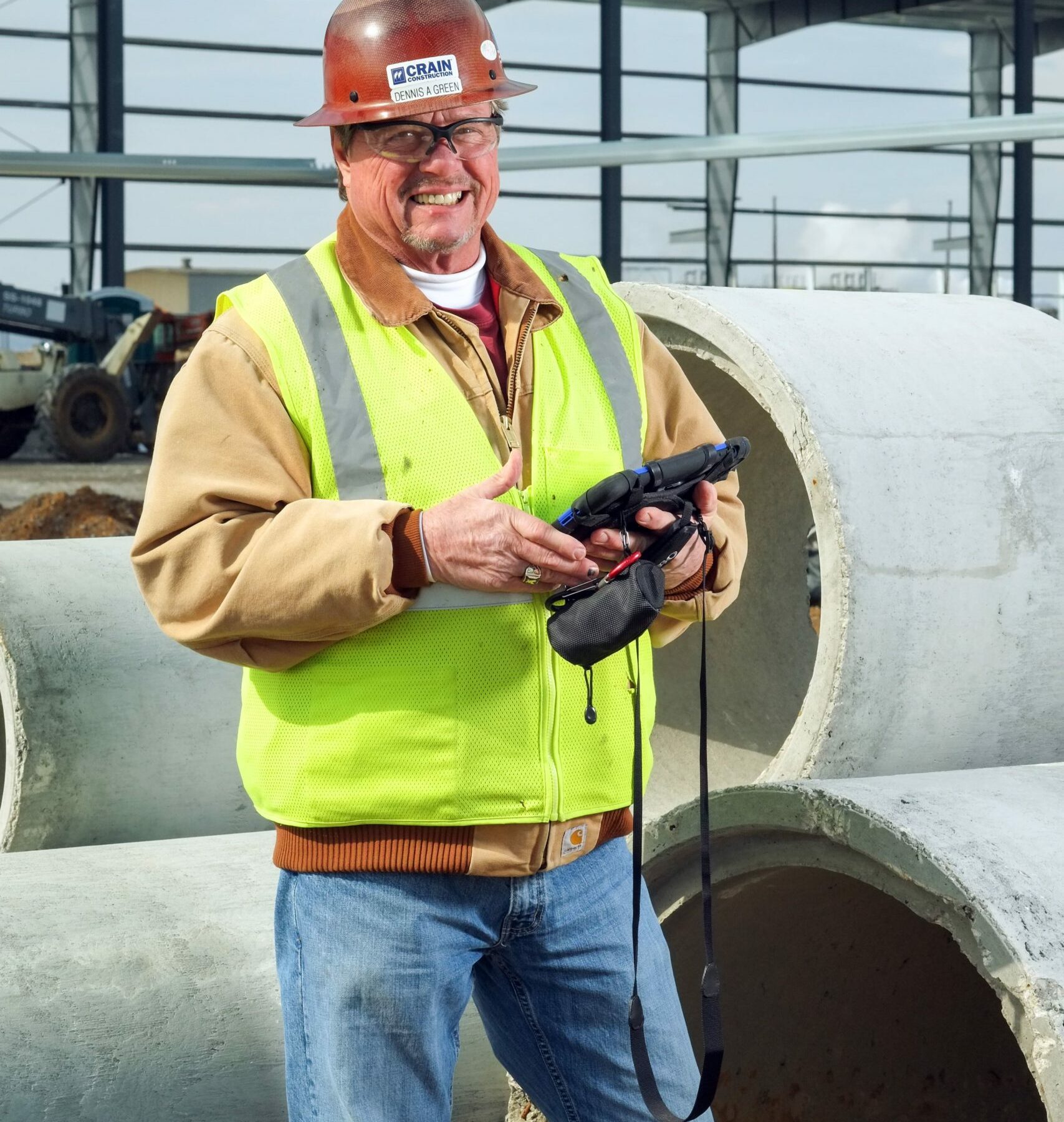 a Crain Construction employee smiling while holding an iPad on a construction site wearing a hard hat and safety vest