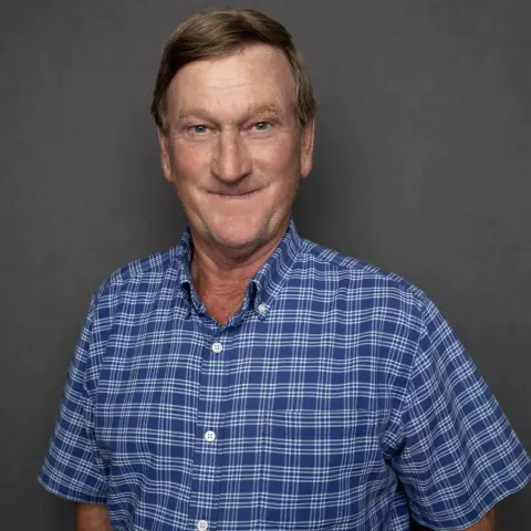 Portrait of JR Howell, a senior superintendent at Crain Construction, in professional attire, smiling and standing against a plain grey background.